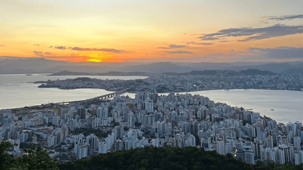 Uma vista panorâmica da cidade de Florianópolis ao entardecer, com o sol se pondo no horizonte, criando tons alaranjados e amarelados no céu. Edifícios altos e densamente agrupados ocupam uma península, ladeada por corpos d'água em ambos os lados, que se estendem até montanhas distantes. Uma ponte pode ser vista conectando a península a outra massa de terra. No primeiro plano, à esquerda e na parte inferior, há vegetação escura.
