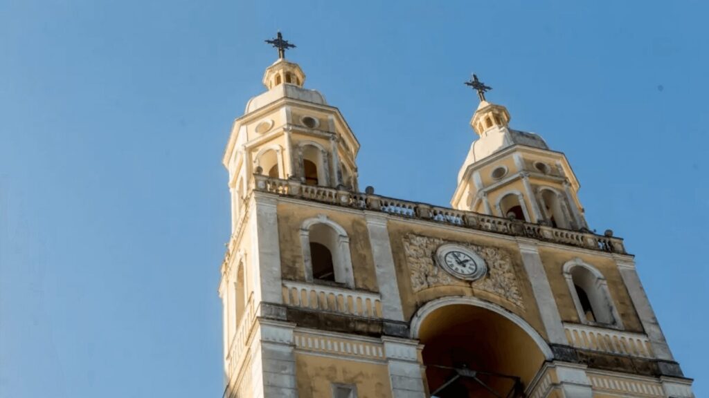 Uma foto de baixo para cima das duas torres sineiras de uma catedral antiga, de cor amarelada com detalhes brancos e ornamentados. Ambas as torres têm cúpulas e cruzes no topo, e a torre da direita é ligeiramente mais alta que a da esquerda. Entre as torres, um relógio está embutido na fachada da catedral. O céu azul claro ocupa a maior parte do fundo da imagem.