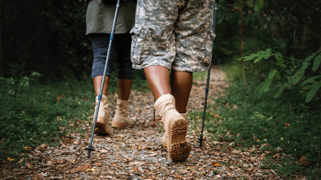 Uma foto de duas pessoas caminhando por uma trilha em uma floresta densa, vistas de trás, dos joelhos para baixo. A pessoa à direita veste uma calça camuflada e botas de cano curto bege, e a pessoa à esquerda veste uma calça escura e botas de cano curto bege. Ambas usam bastões de caminhada. A trilha é coberta por folhas secas e o chão ao redor é coberto por vegetação rasteira.