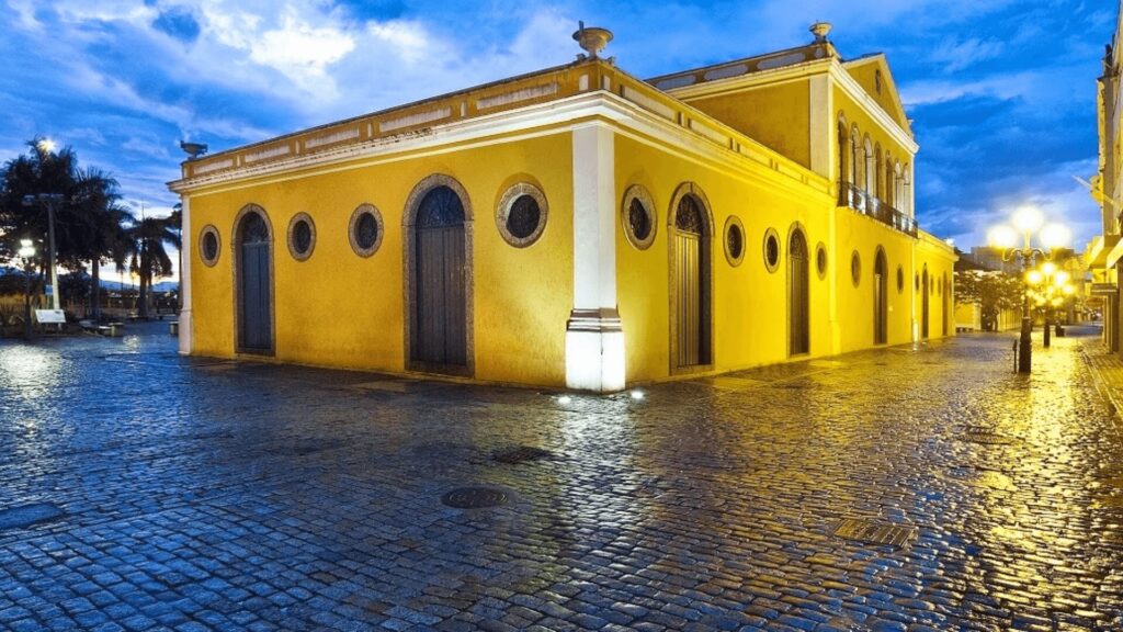 Uma foto noturna de um edifício histórico de cor amarela-mostarda com detalhes brancos, iluminado por luzes artificiais. O edifício possui várias portas e janelas com molduras escuras e algumas janelas redondas. A rua em frente ao edifício é pavimentada com paralelepípedos e parece molhada, refletindo as luzes da rua. No lado direito, há um poste de luz antigo com múltiplas lâmpadas acesas. Ao fundo, o céu é azul escuro com nuvens.