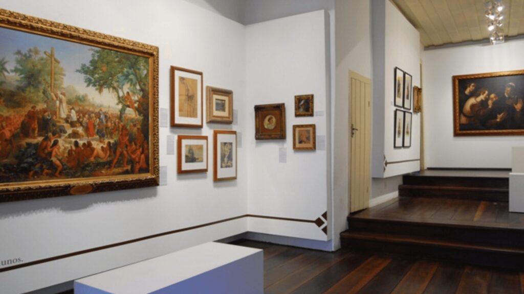 Uma sala de exposição de museu com paredes brancas e piso de madeira escura. À esquerda, uma grande pintura emoldurada em dourado mostra uma cena religiosa com muitas figuras. Várias outras pinturas menores emolduradas estão penduradas na parede adjacente. No centro da sala, há um banco branco. À direita, um lance de escadas de madeira leva a outra área do museu, onde mais pinturas podem ser vistas na parede.