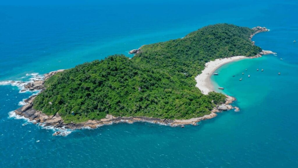 Uma vista aérea de uma ilha verdejante e exuberante, cercada por águas azul-turquesa claras. A ilha possui uma praia de areia branca em um de seus lados, onde alguns barcos estão ancorados. As margens da ilha são rochosas em algumas partes, com vegetação densa cobrindo a maior parte da terra.