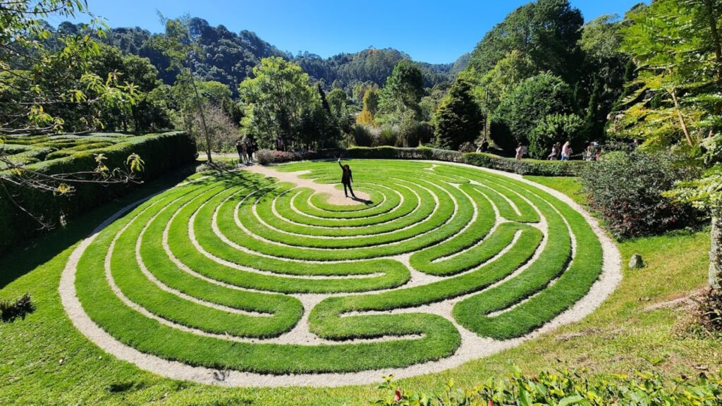 Grande labirinto de meditação circular, com caminhos de grama verde bem aparada e divisórias de cascalho claro, visto de um ângulo ligeiramente elevado em um parque ensolarado. Uma pessoa está de pé no centro do labirinto com um braço erguido. Ao redor e ao fundo, há árvores, vegetação densa, colinas arborizadas e um céu azul limpo. Outras pessoas são visíveis ao longe, fora do labirinto.