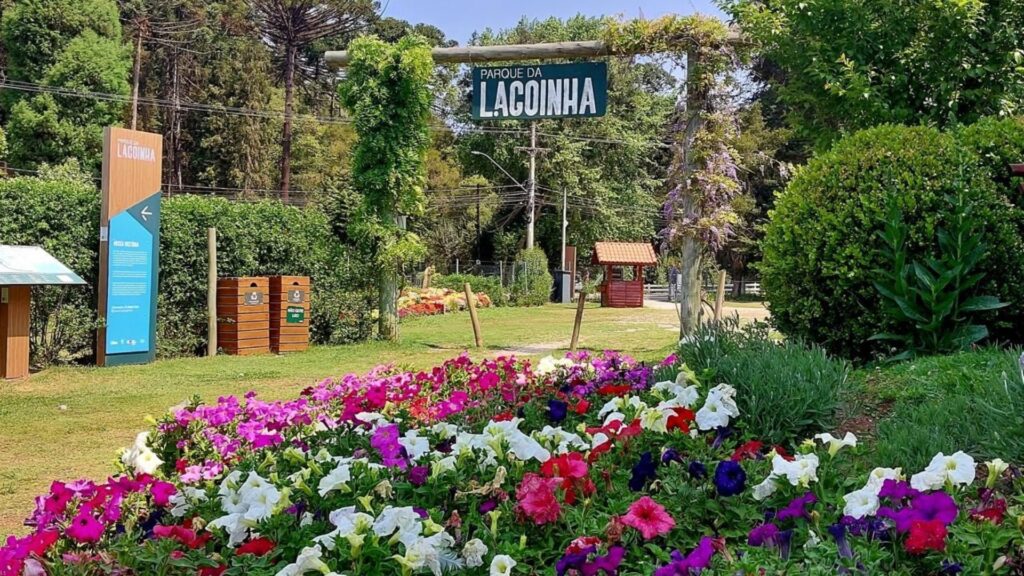 Canteiro vibrante com flores multicoloridas (tons de rosa, roxo, vermelho e branco) em primeiro plano, na entrada do "Parque da Lagoinha". Ao centro, um portal de madeira rústico exibe uma placa verde com o nome do parque em letras brancas. O portal é ladeado por vegetação e árvores altas, incluindo araucárias ao fundo. À esquerda, um painel informativo azul e branco e lixeiras de madeira. Ao fundo à direita, uma pequena estrutura de madeira com telhado. O céu está azul e o dia parece ensolarado.