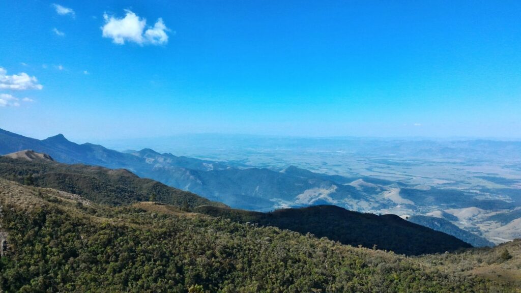 Vista panorâmica de uma vasta paisagem montanhosa sob um céu azul claro com algumas pequenas nuvens brancas. Em primeiro plano, encostas de montanhas cobertas por vegetação densa e verde-escura. As montanhas se estendem em múltiplas camadas, tornando-se mais claras e azuladas à medida que se afastam, até um extenso vale ou planície que se perde no horizonte. A luz do sol ilumina partes da paisagem, enquanto outras permanecem em sombra, realçando o relevo.