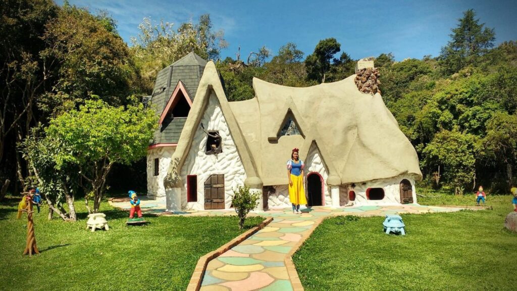 Uma mulher vestida como a personagem Branca de Neve (com vestido azul e amarelo e laço vermelho no cabelo) está em frente a uma casa de estilo rústico e fantasioso, típica de contos de fadas. A casa tem paredes claras, um grande telhado curvo e texturizado de cor bege, janelas pequenas com detalhes em madeira e uma chaminé de pedra. Um caminho feito de pedras coloridas (tons pastel de amarelo, rosa, azul e verde) leva até a porta da casa. No gramado verde em frente, há várias estatuetas pequenas de anões. Ao fundo, árvores densas e um céu azul claro indicam um dia ensolarado em um parque ou floresta.
