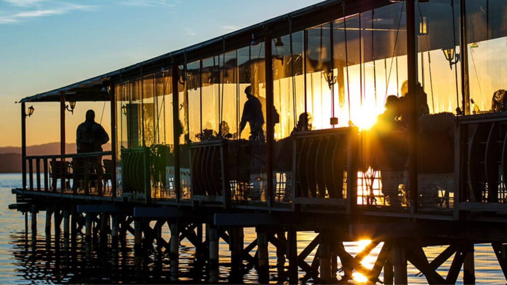 A imagem retrata um restaurante construído sobre palafitas, avançando sobre um corpo de água calmo, como um lago ou baía. A cena é capturada durante o pôr do sol, que cria um efeito de contraluz, transformando as pessoas e objetos em silhuetas escuras contra o brilho intenso do sol.
A estrutura do restaurante é alongada, com um telhado que se estende sobre uma varanda ou área de refeições envidraçada. Grandes painéis de vidro permitem a visualização do interior, onde se percebem silhuetas de pessoas sentadas às mesas e outras em movimento, possivelmente funcionários e clientes. Alguns detalhes da mobília, como mesas e cadeiras, também são visíveis como contornos escuros.
O sol está baixo no horizonte, à direita da imagem, e seu brilho intenso reflete na superfície da água, criando um caminho de luz cintilante. O céu exibe tons quentes, típicos do entardecer, variando do amarelo e laranja perto do sol a um azul mais suave na parte superior.
À esquerda, mais distante, é possível ver a silhueta de montanhas ou uma costa. Os pilares de sustentação do restaurante são visíveis na parte inferior da imagem, fincados na água, com seus reflexos também perceptíveis. A atmosfera geral é serena e pitoresca, destacando a beleza do local ao entardecer.
