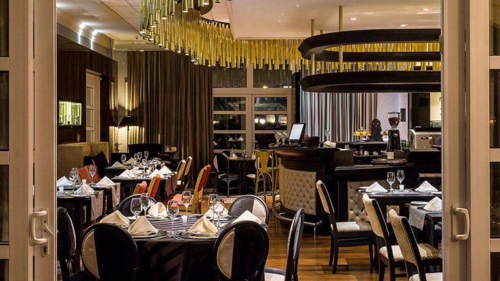 A imagem exibe o interior de um estabelecimento que aparenta ser um restaurante ou bar sofisticado. A foto é tirada a partir de uma porta ou vão, olhando para dentro do salão principal.
O ambiente é elegantemente decorado. No centro e em primeiro plano, há várias mesas redondas cobertas com toalhas escuras, possivelmente pretas. As mesas estão postas com pratos brancos, guardanapos de tecido branco dobrados de forma elaborada, taças de vinho e talheres. As cadeiras ao redor das mesas têm um design clássico e moderno, com encostos arredondados e estofamento que alterna listras pretas e brancas ou um tom creme.
Ao fundo, à direita, destaca-se um balcão de bar com uma estrutura circular escura suspensa sobre ele, que parece ter iluminação embutida. Sobre o balcão, há garrafas, uma máquina de café expresso e outros utensílios de bar. Algumas banquetas altas estão posicionadas junto ao balcão.
A iluminação do ambiente é suave e quente, proveniente de várias fontes, incluindo o que parecem ser tubos dourados ou metálicos pendurados no teto em uma formação decorativa no centro do salão, e luminárias de parede. Grandes janelas ou portas de vidro ao fundo estão cobertas por cortinas longas e escuras, que vão do teto ao chão. As paredes parecem ter painéis de madeira escura ou um revestimento em tons neutros.
O piso não é claramente visível em sua totalidade, mas parece ser de um material polido. A atmosfera geral é de requinte, conforto e um design de interiores cuidadosamente planejado.
