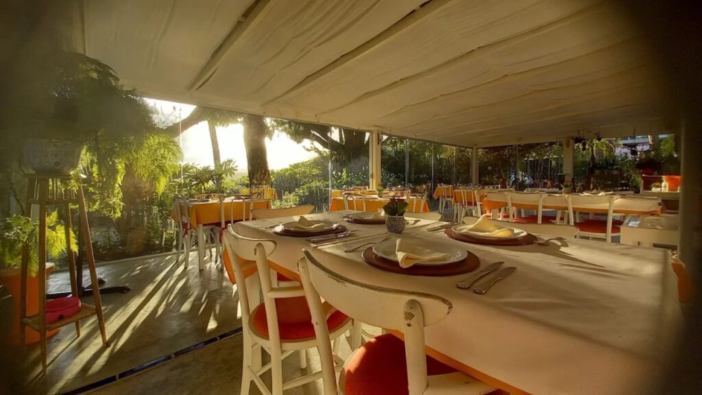 A imagem mostra uma ampla varanda de restaurante, com uma atmosfera clara e iluminada pela luz natural que entra pela lateral esquerda, sugerindo um final de tarde ensolarado ou o nascer do sol.
Em primeiro plano, destaca-se uma grande mesa retangular coberta com uma toalha branca. Sobre ela, estão dispostos pratos rasos de cerâmica ou barro, sousplats, talheres e guardanapos de tecido claro dobrados. As cadeiras ao redor da mesa são brancas com assentos estofados em um tom vibrante de laranja ou vermelho.
Ao fundo, outras mesas menores seguem o mesmo padrão de arrumação, com toalhas de mesa amarelas e cadeiras brancas com assentos coloridos. O espaço é coberto por um teto de lona ou tecido branco retrátil, que difunde a luz. As laterais da varanda parecem ser fechadas por painéis de vidro, permitindo a visualização da vegetação externa.
À esquerda, a luz do sol entra intensamente, criando um brilho e iluminando plantas e folhagens que decoram essa parte do ambiente, incluindo samambaias e outras plantas de vaso. Um grande vaso de cerâmica azul e branco também é visível à esquerda. Ao fundo, através dos vidros e da vegetação, percebe-se um jardim ou área externa com árvores.
O piso da varanda parece ser de cimento queimado ou um material similar, com alguns detalhes em azulejo azul visíveis na borda esquerda. A sensação geral é de um ambiente agradável, arejado e convidativo para uma refeição ao ar livre ou em um espaço semiaberto.