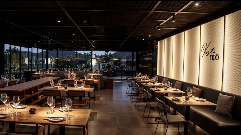 A imagem mostra o interior amplo e moderno de um restaurante durante a noite ou em um ambiente com iluminação controlada. O espaço é caracterizado por uma estética contemporânea e sofisticada.
No centro e à esquerda, diversas mesas de madeira clara, quadradas e retangulares, estão dispostas no salão. As mesas estão postas com pratos, taças de vinho, copos e pequenos arranjos ou luminárias. As cadeiras que acompanham essas mesas são de design simples e escuro. Algumas mesas maiores possuem bancos de madeira compridos em um dos lados.
À direita, ao longo de uma parede, há uma fileira de mesas com um longo sofá estofado (booth) em tom escuro, acompanhado por cadeiras do lado oposto. Esta parede é um destaque visual: é composta por painéis translúcidos ou texturizados que são iluminados por trás, criando um efeito de luz suave e difusa. Sobre esta parede iluminada, está escrito em caligrafia elegante o nome "Vista Nipo".
Ao fundo, grandes janelas de vidro do chão ao teto revelam uma vista externa com vegetação e o que parecem ser luzes de outras edificações ou da paisagem ao anoitecer.
O teto é escuro e possui uma estrutura de grade metálica aparente, de onde pendem pequenas luminárias que fornecem iluminação focal sobre as mesas. A iluminação geral do ambiente é baixa e indireta, criando uma atmosfera íntima e elegante.
O piso é de cor clara e neutra, possivelmente de cimento polido ou um revestimento cerâmico de grande formato. A organização do espaço sugere um local espaçoso e bem planejado. A sensação geral é de um restaurante moderno, com um design de interiores que equilibra elementos industriais e aconchegantes.