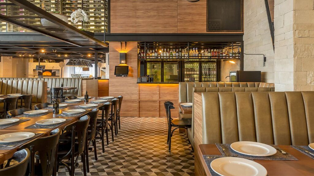 A imagem mostra o interior de um restaurante com um design que mistura elementos rústicos e modernos, criando um ambiente acolhedor.
No primeiro plano e à direita, vemos mesas de madeira escura com pratos brancos e talheres já dispostos. Estas mesas são acompanhadas por longos sofás (booths) com encostos altos e estofamento em couro ou material similar na cor caramelo, com listras verticais. Uma parede de tijolos claros ou pedras é visível à extrema direita.
À esquerda e ao centro, estende-se uma fileira de mesas retangulares de madeira escura, com cadeiras pretas de design simples. As mesas também estão postas com pratos brancos e o que parecem ser garrafas escuras, possivelmente de azeite ou vinho.
O piso é um destaque, com um padrão geométrico em tons de marrom, bege e cinza, formando losangos ou cubos tridimensionais.
Ao fundo, visualiza-se a área do bar, com um balcão revestido em madeira clara e prateleiras de metal preto exibindo diversas garrafas de bebidas. Geladeiras expositoras com mais bebidas são visíveis sob o balcão. Acima do bar e em outras partes do teto, há estruturas metálicas pretas.
Mais ao fundo e à esquerda, é possível entrever o que parece ser um forno a lenha, sugerindo que o local pode ser uma pizzaria ou "forneria". Uma grande estante de vinhos, que vai do teto quase ao chão, repleta de garrafas, é visível na parte superior esquerda, indicando uma boa seleção da bebida. Um arranjo de flores brancas, possivelmente orquídeas, decora uma viga metálica.
A iluminação é uma combinação de luzes embutidas e pendentes, criando um ambiente com luminosidade agradável e focada nas mesas e no bar. As paredes alternam entre revestimento de madeira clara e a alvenaria de pedra, contribuindo para a atmosfera rústica e elegante.