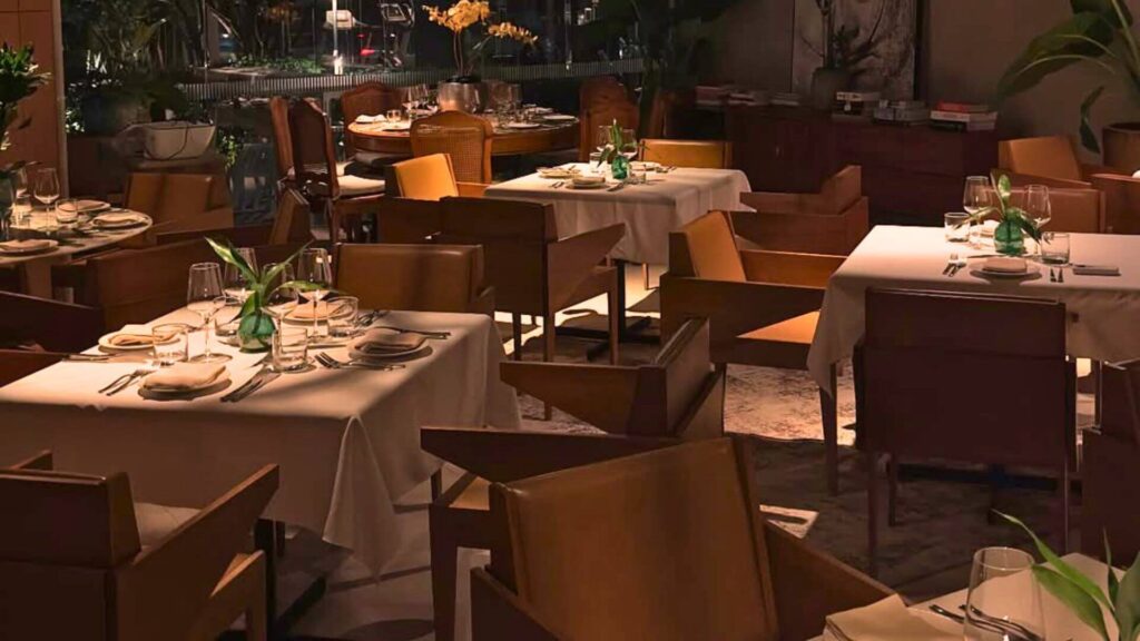 Visão interna de um restaurante elegante e com iluminação suave. Em primeiro plano, mesas quadradas estão postas com toalhas brancas, pratos, talheres, taças e pequenos arranjos de plantas verdes. As cadeiras são de madeira com assentos e encostos estofados em tom claro, possivelmente bege ou caramelo. Ao fundo, mais mesas seguem o mesmo padrão, e podem-se ver plantas maiores, armários de madeira e o que parece ser uma área de bar ou balcão de serviço. A atmosfera geral é acolhedora e sofisticada, com foco nos tons terrosos e na iluminação indireta.