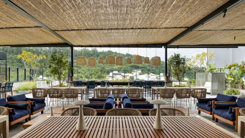 Amplo pátio ou terraço de restaurante com um teto distintivo feito de fileiras de bambu ou palha, sustentado por uma estrutura metálica escura. Em primeiro plano, uma grande mesa de madeira ripada clara com dois pequenos candeeiros de mesa modernos, de cúpula branca e base fina. Flanqueando esta mesa, há conjuntos de sofás azuis escuros com almofadas claras e estrutura de madeira. Mais ao fundo, diversas mesas de jantar, algumas quadradas e outras retangulares, com cadeiras de vime ou material natural trançado e almofadas claras. Sobre a área de jantar central, pendem várias luminárias grandes em forma de cesto, feitas de material natural trançado. O espaço é aberto nas laterais, com um guarda-corpo de vidro, oferecendo uma vista para uma paisagem com vegetação densa e algumas construções ao longe, sob um céu nublado. O ambiente é arejado, iluminado e combina elementos naturais e rústicos com um design contemporâneo.