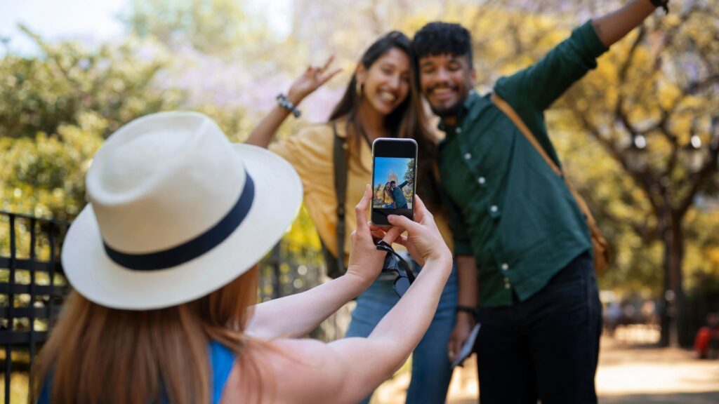 A imagem captura uma cena ao ar livre onde uma pessoa de costas, em primeiro plano, tira uma foto com um celular de um casal que posa para a câmera.
A pessoa em primeiro plano tem cabelos ruivos visíveis e usa um chapéu de palha claro com uma faixa preta. O celular que ela segura está em modo retrato, e a tela mostra a imagem do casal que está sendo fotografado.
O casal, em segundo plano e ligeiramente desfocado, sorri e faz poses para a foto. A mulher, à esquerda, tem cabelos longos e escuros, veste uma blusa amarela e faz um sinal de "paz" com a mão. O homem, à direita, tem cabelos curtos e escuros, barba, veste uma camisa verde-escura e tem um braço estendido para o lado. Ambos parecem estar em um ambiente com vegetação ao fundo, possivelmente um parque ou área verde. A luz sugere um dia claro e ensolarado.