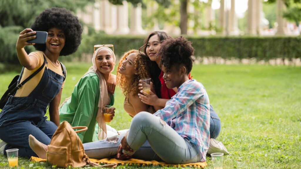 A imagem mostra cinco jovens mulheres diversas, aparentemente amigas, sentadas em um gramado verde durante o dia, desfrutando de um momento de lazer.
No canto esquerdo, uma mulher com cabelo crespo volumoso está segurando um celular na altura do rosto, aparentemente tirando uma selfie ou gravando um vídeo, e sorri amplamente para a câmera. Ela veste um macacão jeans e uma camiseta de cor clara por baixo. Ao seu lado, uma mulher de touca clara e óculos de sol na cabeça sorri, vestindo uma camisa verde-clara e um hijab que cobre parte do cabelo.
No centro da imagem, outra mulher com cabelos cacheados e volumosos, provavelmente com uma pele mais escura, ri com a cabeça inclinada para trás e para o lado, segurando um copo transparente com uma bebida cor de âmbar. Próxima a ela, uma mulher de cabelos escuros e lisos, de origem asiática, sorri e se inclina levemente.
Mais à direita, uma mulher com cabelos cacheados e volumosos, também de pele mais escura, está sentada de lado, com as pernas cruzadas, vestindo uma camisa xadrez azul e branca e calças jeans. Ela segura um copo com uma bebida similar e parece sorrir.
Há copos com bebidas espalhados pelo gramado e uma bolsa marrom clara ao lado da mulher que está tirando a foto. O fundo é composto por árvores e folhagens, sugerindo um parque ou área verde. A atmosfera geral é de alegria, amizade e descontração.