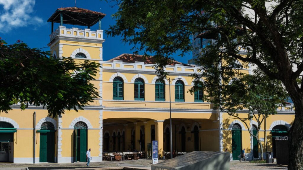 A imagem apresenta o Mercado Público de Florianópolis, um edifício histórico e arquitetonicamente interessante, com fachada em tons de amarelo claro e detalhes em branco. O prédio possui uma estrutura imponente, com múltiplos arcos e janelas. No lado esquerdo, destaca-se uma torre que se eleva acima do restante da construção, com uma área superior que parece ser um mirante ou observatório, coberta por um telhado de telhas avermelhadas.