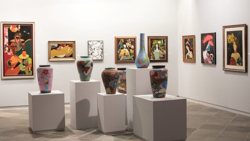 A imagem mostra o interior de uma galeria de arte, provavelmente o Museu de Arte de Santa Catarina, com paredes brancas e piso cinza claro, criando um ambiente minimalista que realça as obras expostas.