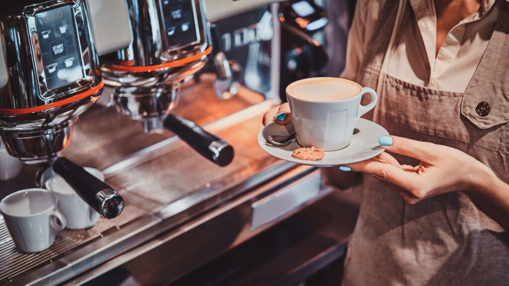 A imagem foca em um momento em uma cafeteria, com uma pessoa (provavelmente um barista) segurando uma xícara de café recém-preparado. A xícara branca, com um pires e uma pequena colher, contém uma bebida com latte art delicado na superfície. Ao lado da xícara, no pires, há um pequeno biscoito.