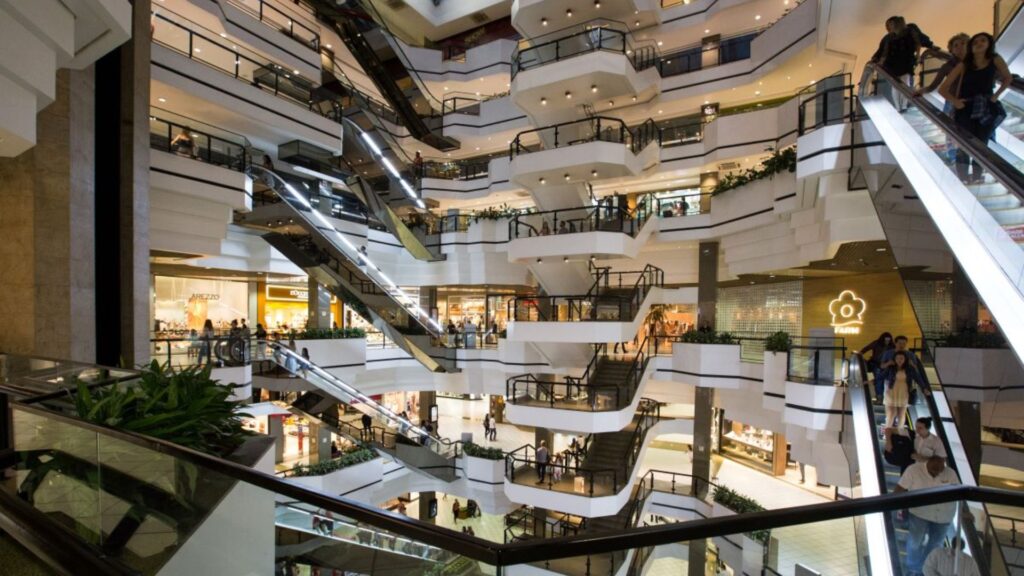 A imagem mostra o interior de um shopping center moderno e espaçoso, caracterizado por múltiplos níveis interligados por escadas rolantes e escadas fixas. O design do ambiente é dominado por tons claros, com paredes e estruturas em branco ou bege claro, complementadas por detalhes em preto, como os corrimãos das escadas e grades de proteção.