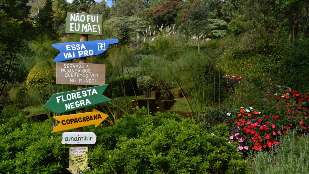 Fotografia de um jardim vibrante (possivelmente Amantikir) com um poste de sinalização central feito de madeira. No poste, diversas placas em formato de seta, pintadas em cores como azul, verde e amarelo, além de placas retangulares em madeira natural, indicam direções ou frases humorísticas e inspiradoras.