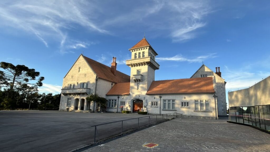 Fachada do Palácio Boa Vista, uma construção imponente com paredes claras, telhados de terracota e uma torre central, sob um céu azul com nuvens. Um pátio pavimentado está em primeiro plano e a copa de uma araucária é visível à esquerda.