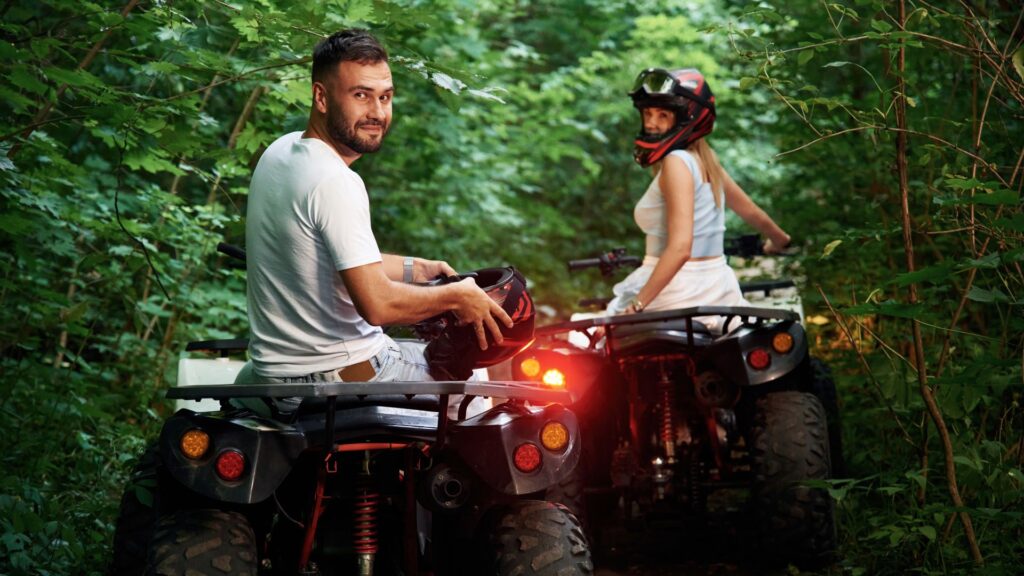 Casal em dois quadriciclos em uma trilha de floresta. O homem à frente, sem capacete e sorrindo para a câmera, segura um capacete. A mulher atrás usa capacete e olha por cima do ombro.