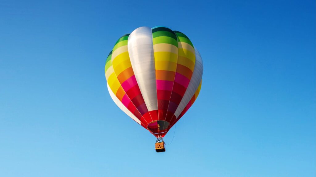 Balão de ar quente multicolorido, com listras verticais vibrantes em tons de vermelho, laranja, amarelo, verde, azul, rosa e branco, flutuando em um céu azul claro e sem nuvens. Uma cesta de vime marrom está suspensa abaixo.