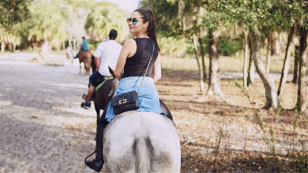 Mulher jovem montada em um cavalo de pelagem clara, vista de costas, olhando por cima do ombro para a câmera. Ela usa óculos de sol, regata preta, saia jeans e uma bolsa preta transpassada. Outros cavaleiros ao fundo em uma trilha arborizada.