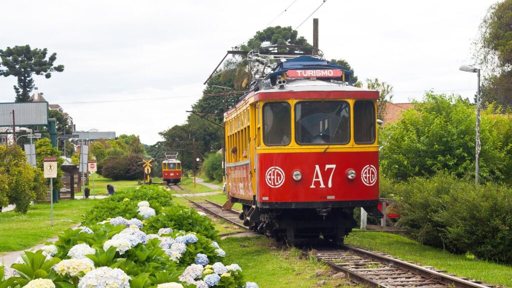 Bonde elétrico vermelho e amarelo, identificado como 'A7' e com placa 'TURISMO', em trilhos cercados por vegetação e um canteiro de hortênsias azuis e brancas em primeiro plano. Outro bonde similar ao fundo, sob céu nublado, em um cenário que remete a Campos do Jordão.