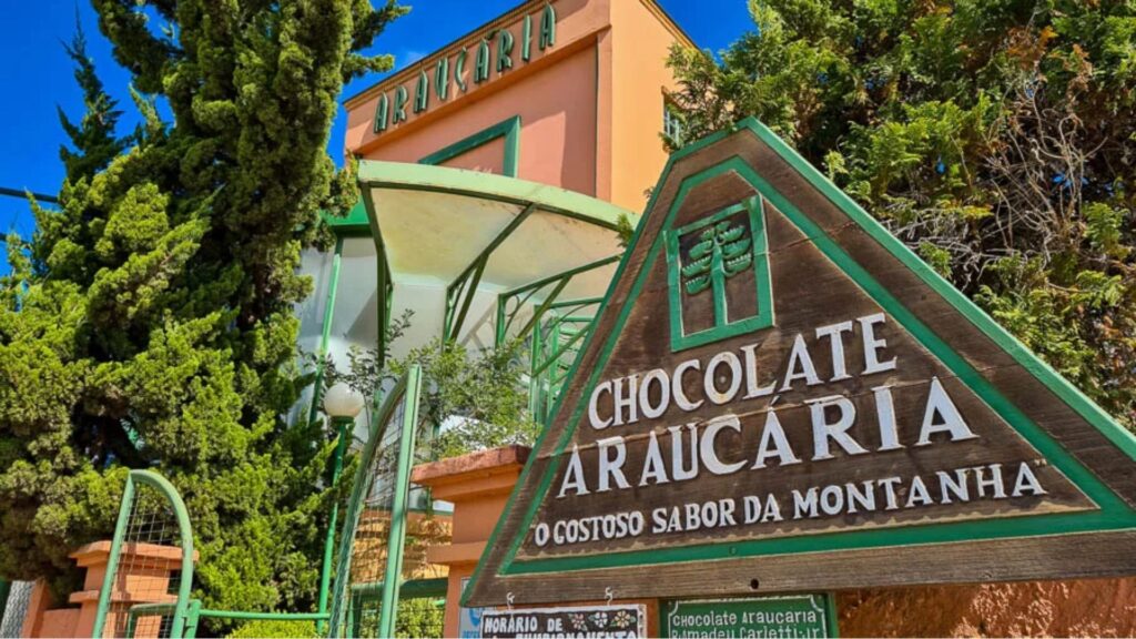 Placa de madeira triangular em primeiro plano indicando 'CHOCOLATE ARAUCÁRIA - O GOSTOSO SABOR DA MONTANHA'. Ao fundo, um edifício cor pêssego com o nome 'ARAUCÁRIA' na fachada, cercado por árvores altas sob um céu azul. Provável entrada de uma fábrica ou loja de chocolates em Campos do Jordão.