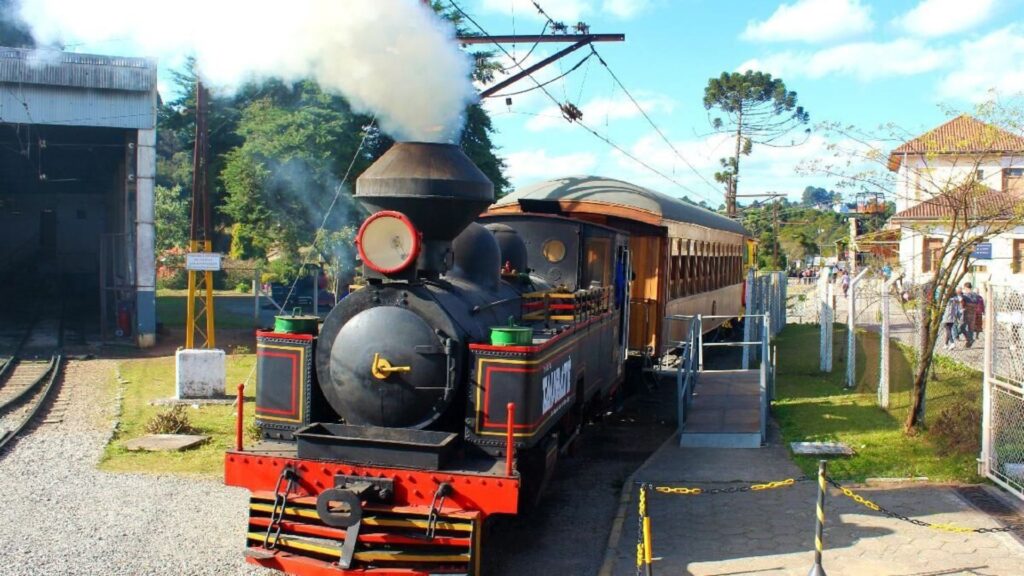 Locomotiva a vapor preta (Maria Fumaça) com detalhes em vermelho e dourado, parada em uma estação, expelindo fumaça branca. Um vagão de passageiros amarelo está acoplado. Pessoas na plataforma e árvores ao fundo, sob céu azul.