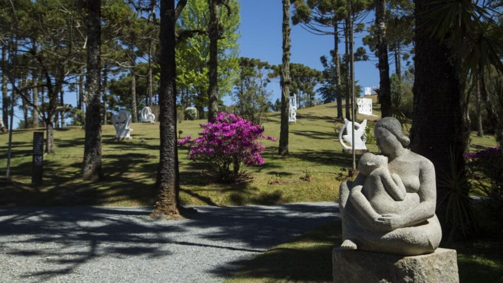 Escultura de pedra de uma mulher com uma criança ao colo em primeiro plano à direita, em um parque arborizado. Ao fundo, um arbusto com flores rosa-púrpura vibrantes, outras esculturas claras e altas araucárias sob um céu azul.