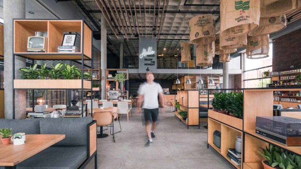 A imagem mostra o interior de uma cafeteria ou restaurante espaçoso e moderno com um design industrial e rústico. A foto é tirada de uma perspectiva ampla, capturando vários detalhes do ambiente.
No centro da imagem, um homem com uma camiseta branca e shorts escuros caminha em direção à câmera; seu movimento é capturado com um leve desfoque. Acima dele, pendurado no teto alto, há um letreiro de neon que diz "Hello! Enjoy the moment" ("Olá! Aproveite o momento").
O teto é de concreto com uma textura quadriculada e tubulações expostas. Grandes luminárias pendentes, feitas de sacos de juta de café com a inscrição "Café do Brasil", iluminam o espaço, adicionando um toque temático e rústico.
As paredes e o piso são de cimento queimado. O mobiliário é composto por grandes estantes de metal preto com prateleiras de madeira clara, que dividem o ambiente e servem como decoração. As estantes contêm vários objetos, como plantas, livros, uma televisão antiga de tubo e outros itens decorativos.