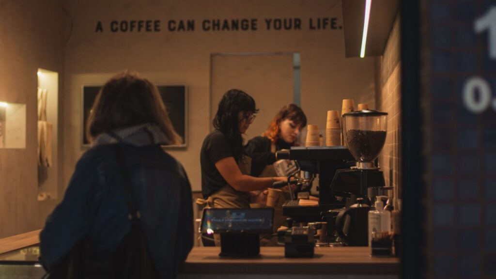 A imagem mostra o interior de uma cafeteria moderna com iluminação quente e ambiente intimista. A cena é vista por cima do ombro de uma cliente que está em primeiro plano, de costas para a câmera. Ela tem cabelo curto e castanho e veste uma jaqueta jeans.
A cliente está em frente a um balcão de madeira, onde duas baristas estão trabalhando. Uma barista, com cabelo escuro e na altura dos ombros, está concentrada operando uma grande máquina de café expresso. Ao lado dela, outra barista com cabelo avermelhado e óculos a observa.
No balcão, além da máquina de expresso, há um moedor de café cheio de grãos, pilhas de copos de papel descartáveis e um terminal de pagamento.
Na parede do fundo, logo acima da área de trabalho das baristas, a frase em inglês "A COFFEE CAN CHANGE YOUR LIFE." (Um café pode mudar a sua vida.) está escrita em letras maiúsculas escuras. A iluminação geral é suave e focada na área do balcão, criando uma atmosfera calma e acolhedora.