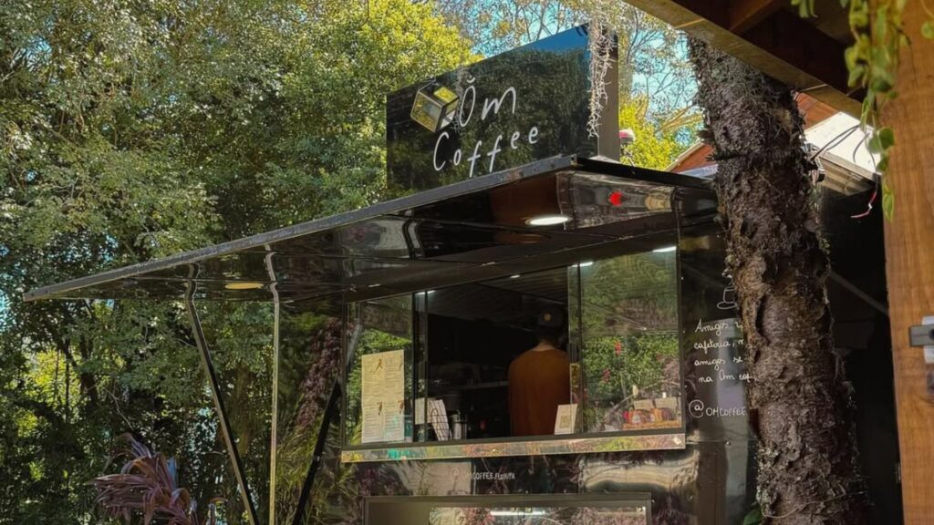 A imagem mostra um quiosque ou food truck de café, moderno e elegante, posicionado em uma área externa cercada por natureza. O quiosque é predominantemente preto, com uma superfície brilhante que reflete a luz.
Uma grande janela de atendimento está aberta, com um toldo preto largo estendido sobre ela para fornecer sombra. No topo do quiosque, uma placa preta exibe um logotipo de um cubo amarelo estilizado ao lado do nome "Om Coffee", escrito em uma fonte branca que imita a caligrafia.
Dentro do quiosque, é possível ver a silhueta de um barista de costas, vestindo uma blusa de cor marrom ou mostarda. No balcão, há um menu e outros itens de trabalho. No vidro, à direita da janela, há um texto escrito em branco e o perfil do Instagram "@OMCOFFEE".
O fundo da imagem é preenchido por árvores altas e uma folhagem verde e densa, indicando que o quiosque está localizado em um parque, jardim ou espaço arborizado. À direita da foto, um tronco de árvore robusto com plantas penduradas se integra à cena. A iluminação é natural e diurna, criando um contraste interessante entre o design moderno do quiosque e o ambiente natural e tranquilo.