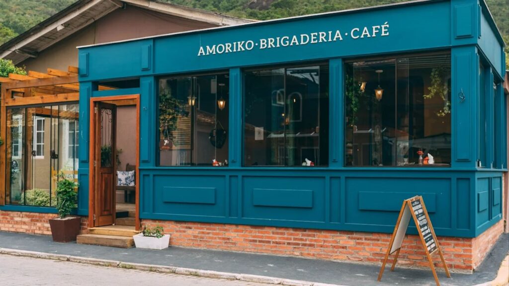 A imagem exibe a fachada de uma cafeteria charmosa chamada "AMORIKO · BRIGADERIA · CAFÉ". O nome está escrito em letras brancas, elegantes e em caixa alta, em um painel que corre acima de grandes janelas.
A arquitetura da loja é o destaque principal. A fachada é pintada em um tom forte e sofisticado de azul-petróleo. A parte inferior da parede é de tijolos vermelhos aparentes, assentada sobre a calçada. A loja possui três grandes janelas de vidro que permitem ver o interior aconchegante, com luminárias pendentes acesas.
A porta de entrada, com detalhes em madeira e vidro, fica um pouco recuada à esquerda. Ao lado da porta, há um pequeno vaso de plantas retangular e branco no chão. À esquerda da entrada principal, há uma extensão da loja que parece uma varanda ou pátio fechado com grandes painéis de vidro e uma estrutura de pérgola de madeira no topo.
Na calçada, do lado direito da imagem, um cavalete de madeira (placa tipo sanduíche) exibe um menu ou informações para os clientes. Ao fundo, pode-se ver o telhado de uma casa vizinha e uma montanha coberta por vegetação verde, sob um céu claro, indicando que o local é possivelmente uma cidade pequena ou uma área de serra. O aspecto geral é de um estabelecimento convidativo, limpo e bem decorado.