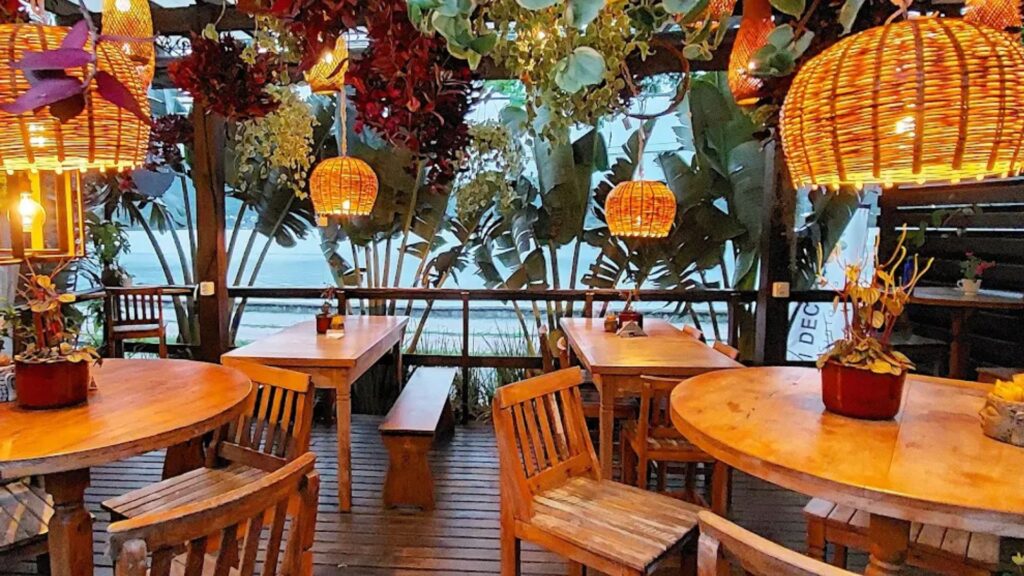 A imagem captura o ambiente de um restaurante ou café com uma decoração intensamente rústica e tropical. O local parece ser uma varanda ou um pátio coberto, com o piso e todos os móveis feitos de madeira em tons naturais.
O espaço está mobiliado com diversas mesas, algumas redondas e outras retangulares, acompanhadas por cadeiras e bancos de madeira com um design simples. O ambiente é iluminado por múltiplas luminárias pendentes, que são um ponto focal da decoração. Suas cúpulas são feitas de vime ou outra fibra natural trançada, em formato de cesto, e emitem uma luz muito quente, de cor amarela-alaranjada, criando uma atmosfera extremamente acolhedora e íntima.
O teto é uma estrutura de madeira tipo pérgola, da qual pendem não apenas as luzes, mas também uma enorme quantidade de plantas e folhagens. Há uma mistura densa de folhas verdes, creme e, proeminentemente, de um tom vermelho-escuro ou vinho, que criam um tipo de dossel sobre as mesas.
Ao fundo, através das aberturas da varanda, pode-se ver o exterior com grandes folhas de plantas tropicais, como bananeiras. A iluminação sugere ser o início da noite. O conjunto transmite uma sensação de refúgio natural, boêmio e muito aconchegante.