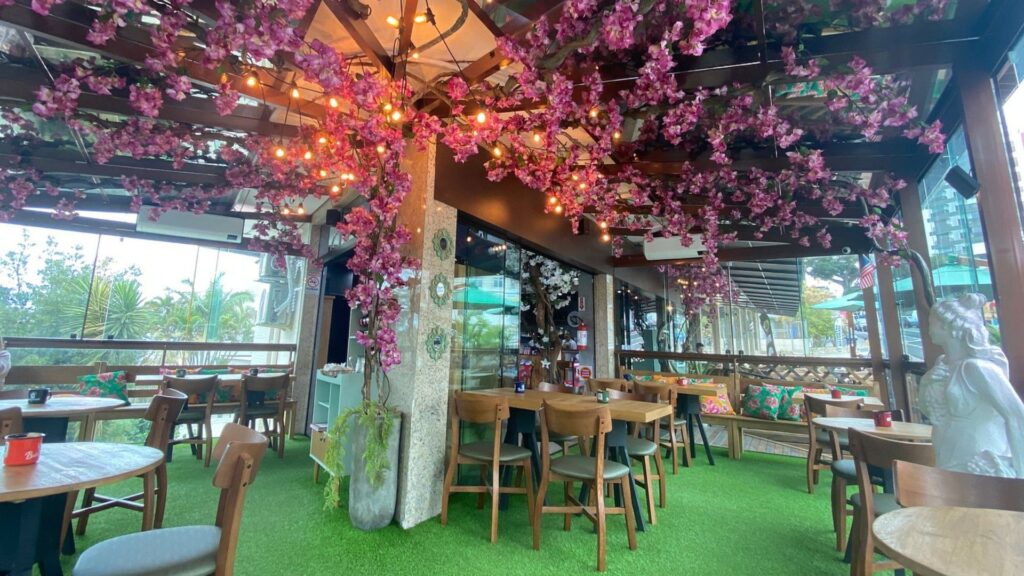 A imagem mostra o interior de um restaurante ou café com uma decoração muito elaborada e fantasiosa, que cria a sensação de se estar em um jardim encantado.
O elemento mais marcante é o teto, que é coberto por uma pérgola de madeira escura. Entrelaçada por toda a estrutura, há uma copa densa de galhos e flores artificiais em tons de rosa e roxo, imitando cerejeiras em flor. Misturadas a essa folhagem, há cordões de luzes com lâmpadas de filamento, que emitem um brilho quente e dourado, conferindo um ar mágico ao ambiente.
O chão do estabelecimento é todo forrado com grama sintética de um verde bem vivo, reforçando a temática de jardim. Os móveis são de madeira em estilo contemporâneo, com mesas quadradas e redondas, e cadeiras com assentos escuros.
Um pilar central é revestido com uma textura clara e decorado com pequenos azulejos. No lado direito da imagem, uma estátua branca de uma figura feminina pequena adiciona ao charme do local. O espaço parece ser uma varanda fechada com vidro, pois ao fundo é possível ver o ambiente externo com árvores, incluindo uma palmeira. A atmosfera geral é romântica, lúdica e muito "instagramável".