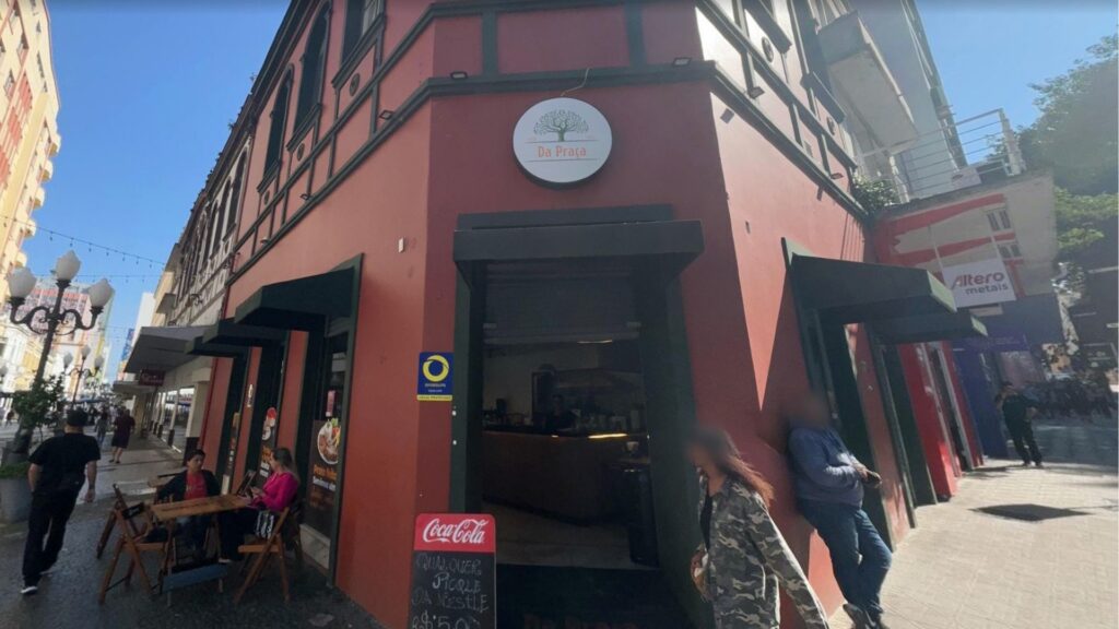 Fachada do café "Da Praça" em um prédio de esquina, de cor terracota, localizado em uma rua movimentada de pedestres. Pessoas estão sentadas em mesas na calçada e caminham pela rua sob um céu azul e ensolarado.
