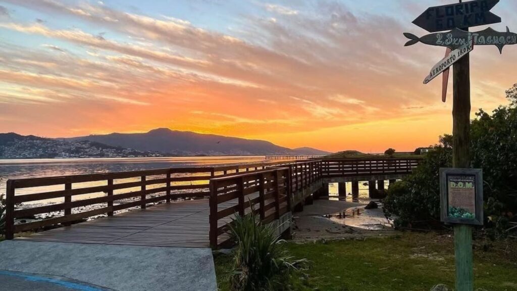 A imagem retrata um pôr do sol espetacular sobre um corpo d'água, provavelmente um estuário ou uma grande baía, com uma paisagem de montanhas ao fundo. O céu está tingido de tons vibrantes de laranja, amarelo e rosa, indicando o crepúsculo.