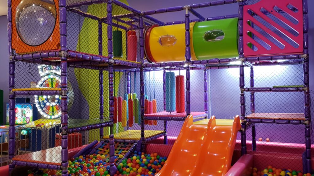 A imagem mostra uma grande e colorida estrutura de playground interno, projetada para crianças, com vários níveis e atividades.