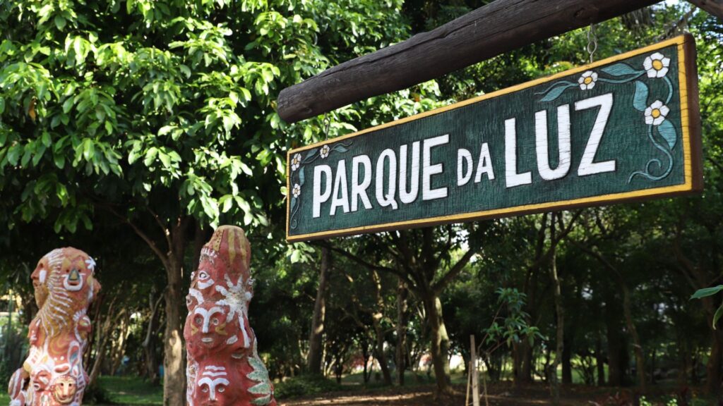 A imagem é um close-up de uma placa de madeira rústica, escura, pendurada por um suporte que parece ser um tronco de árvore horizontal, em um ambiente de parque ou jardim.