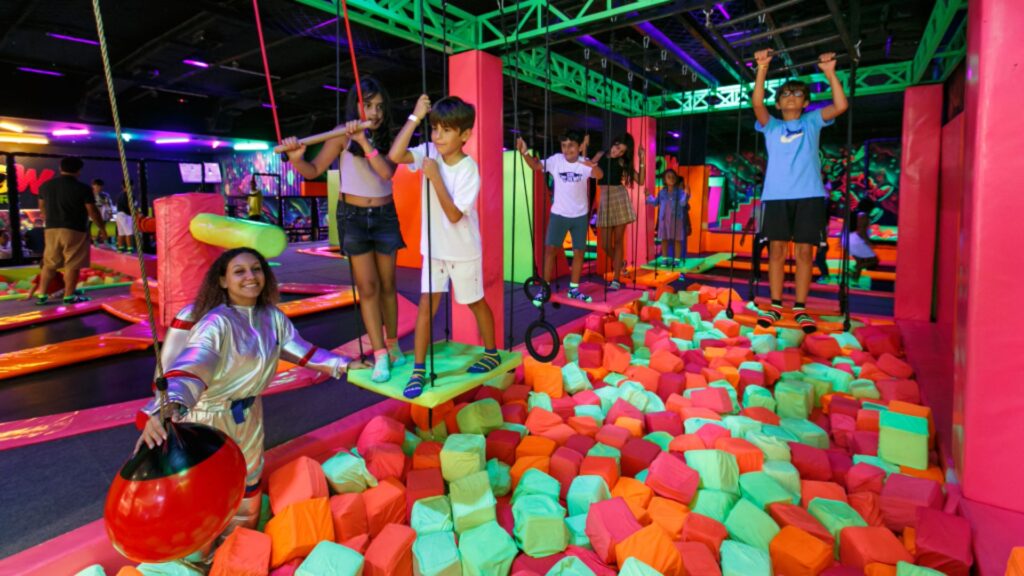 A imagem retrata um parque de trampolins ou playground indoor vibrante e cheio de energia, com cores neon dominantes, principalmente rosa choque e verde limão.