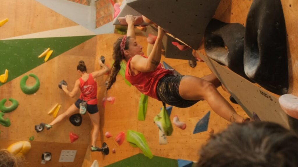 A imagem captura uma cena em uma parede de escalada indoor, ou boulder, onde duas mulheres estão ativamente praticando.