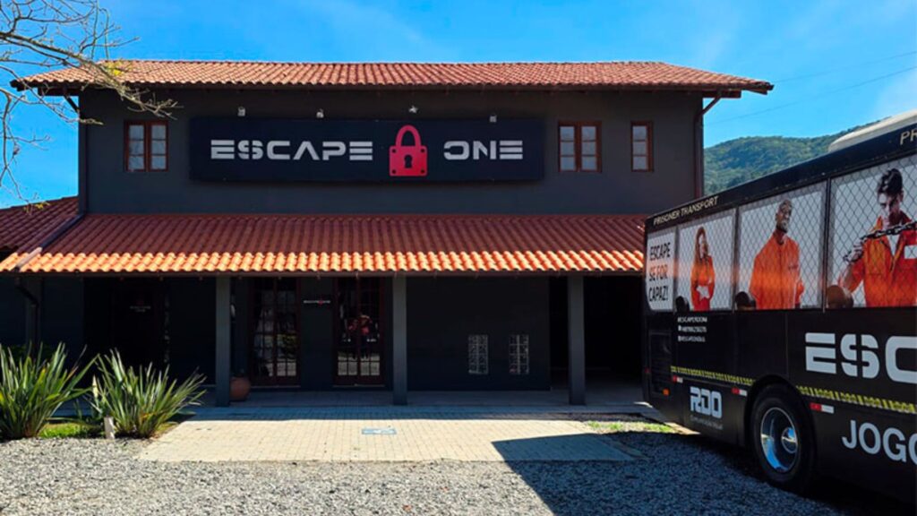 A imagem mostra a fachada de um edifício que abriga um jogo de "escape room", com um ônibus temático estacionado à direita.
O edifício tem dois andares, com paredes em um tom escuro de cinza ou marrom e um telhado de telhas cerâmicas avermelhadas. No andar superior, há três janelas retangulares com molduras escuras. No centro da fachada superior, um letreiro luminoso exibe o nome "ESCAPE ONE" em letras brancas estilizadas, com um ícone de cadeado vermelho entre as palavras "ESCAPE" e "ONE".
A entrada principal do edifício, no térreo, é marcada por uma porta de vidro. Há vasos de plantas na entrada. Na frente do prédio, há um pátio de cascalho claro e um caminho de tijolos ou pedras.
À direita da imagem, está estacionado um ônibus grande e escuro, com adesivos temáticos que promovem o jogo de escape room. Nos adesivos, é possível ver imagens de pessoas com uniformes laranja, semelhantes a prisioneiros, e o texto "PRISONER TRANSPORT" e "ESCAPE SE FOR CAPAZ".
A vegetação baixa é visível em frente ao edifício, e ao fundo, à direita, há uma área montanhosa com árvores. O céu está claro e azul, sugerindo um dia ensolarado.