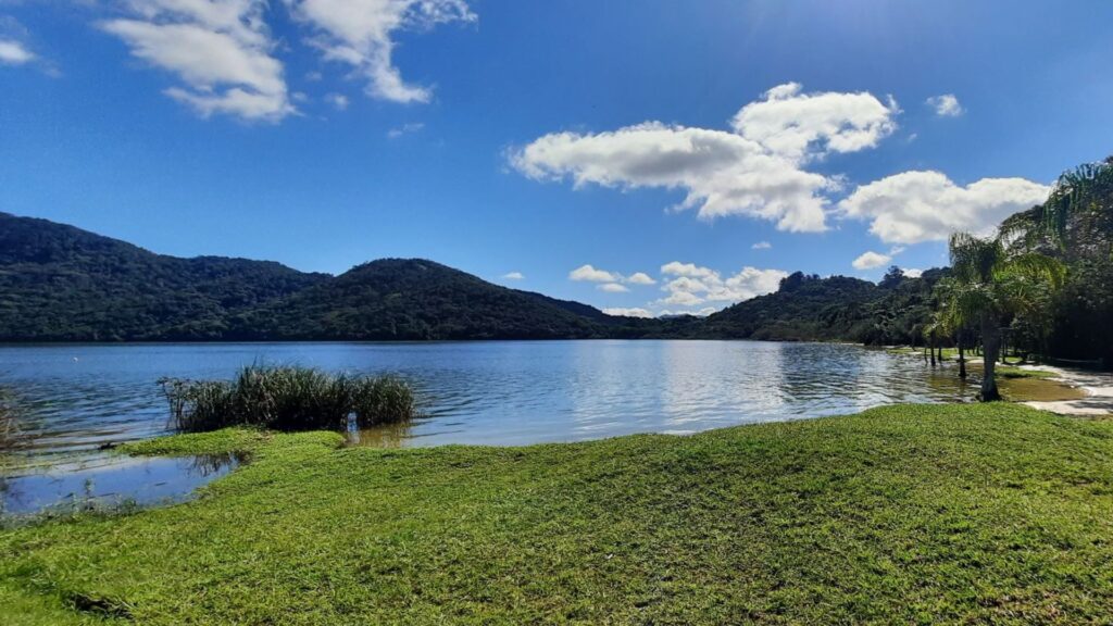 A imagem mostra um grande corpo d'água, possivelmente uma lagoa ou um lago, com uma margem de areia ou terra em primeiro plano. A água reflete o céu e a vegetação ao redor, criando uma superfície relativamente calma.