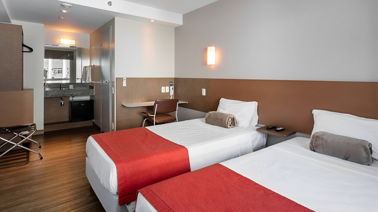 Hotel econômico em Curitiba Um quarto de hotel do Go Inn Curitiba com duas camas de solteiro. As camas têm lençóis brancos e mantas vermelhas aos pés. Entre elas, uma pequena mesa de apoio com controle remoto e telefone. Há uma cabeceira em tom de madeira e uma luminária de parede. Ao fundo, à esquerda, é visível uma área de cozinha compacta ou copa, com uma pia de granito, espelho e um frigobar. À direita, uma pequena escrivaninha e uma cadeira. O quarto possui piso de madeira clara e paredes em tons neutros, transmitindo uma sensação de modernidade e funcionalidade.