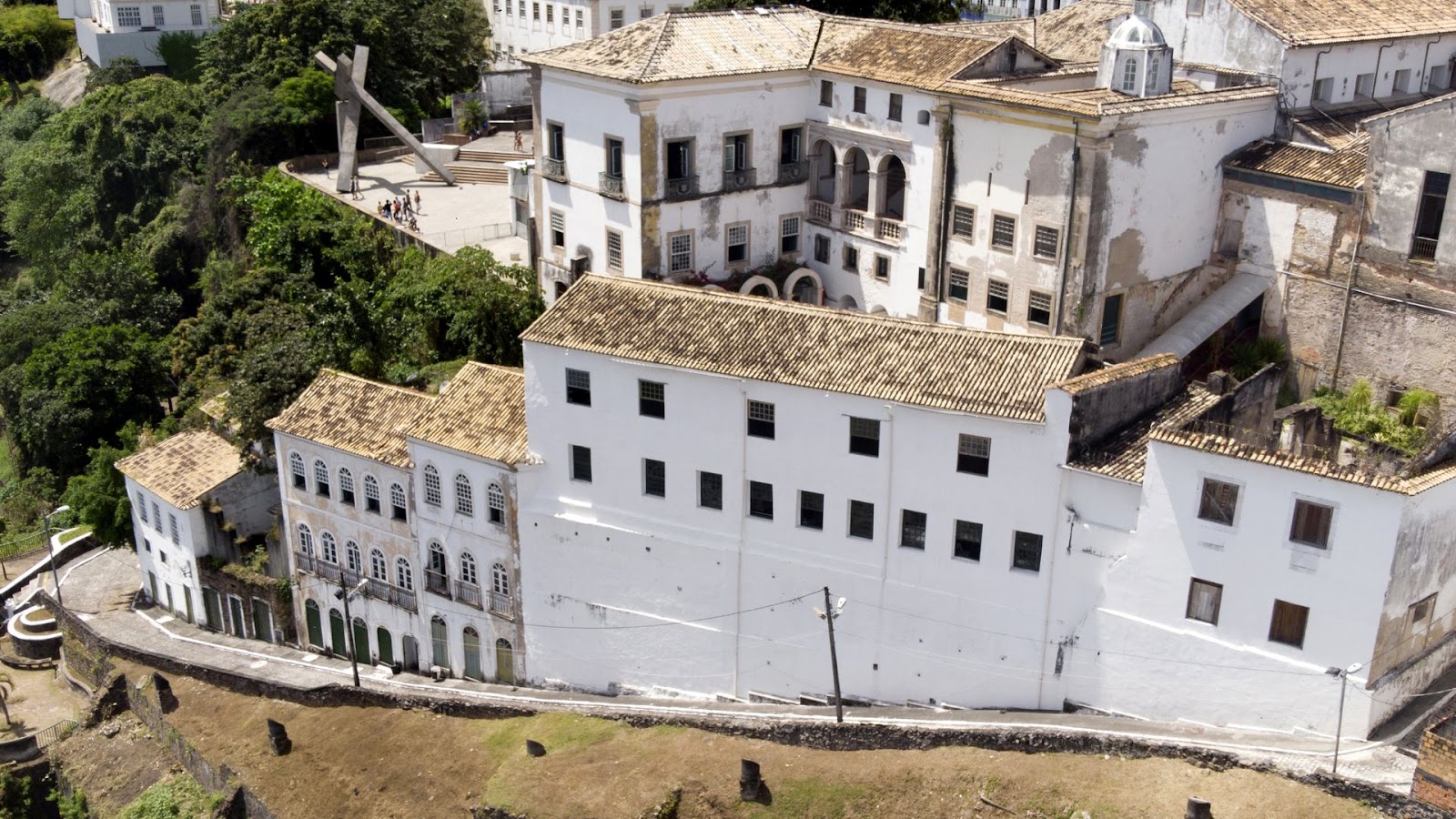 Arquitetura em Salvador Uma vista aérea ou de ângulo elevado da Ladeira da Misericórdia em Salvador, Bahia. A imagem mostra um conjunto de edifícios históricos brancos com telhados de telhas cerâmicas em tons terrosos, dispostos em uma encosta íngreme. O prédio maior e mais proeminente no topo possui várias janelas escuras e varandas, além de uma cúpula. Abaixo dele, uma série de construções menores se estendem ao longo da ladeira, que é ladeada por muros de pedra e vegetação exuberante. No canto superior esquerdo, é possível ver parte de uma praça com uma estrutura artística em forma de cruz.