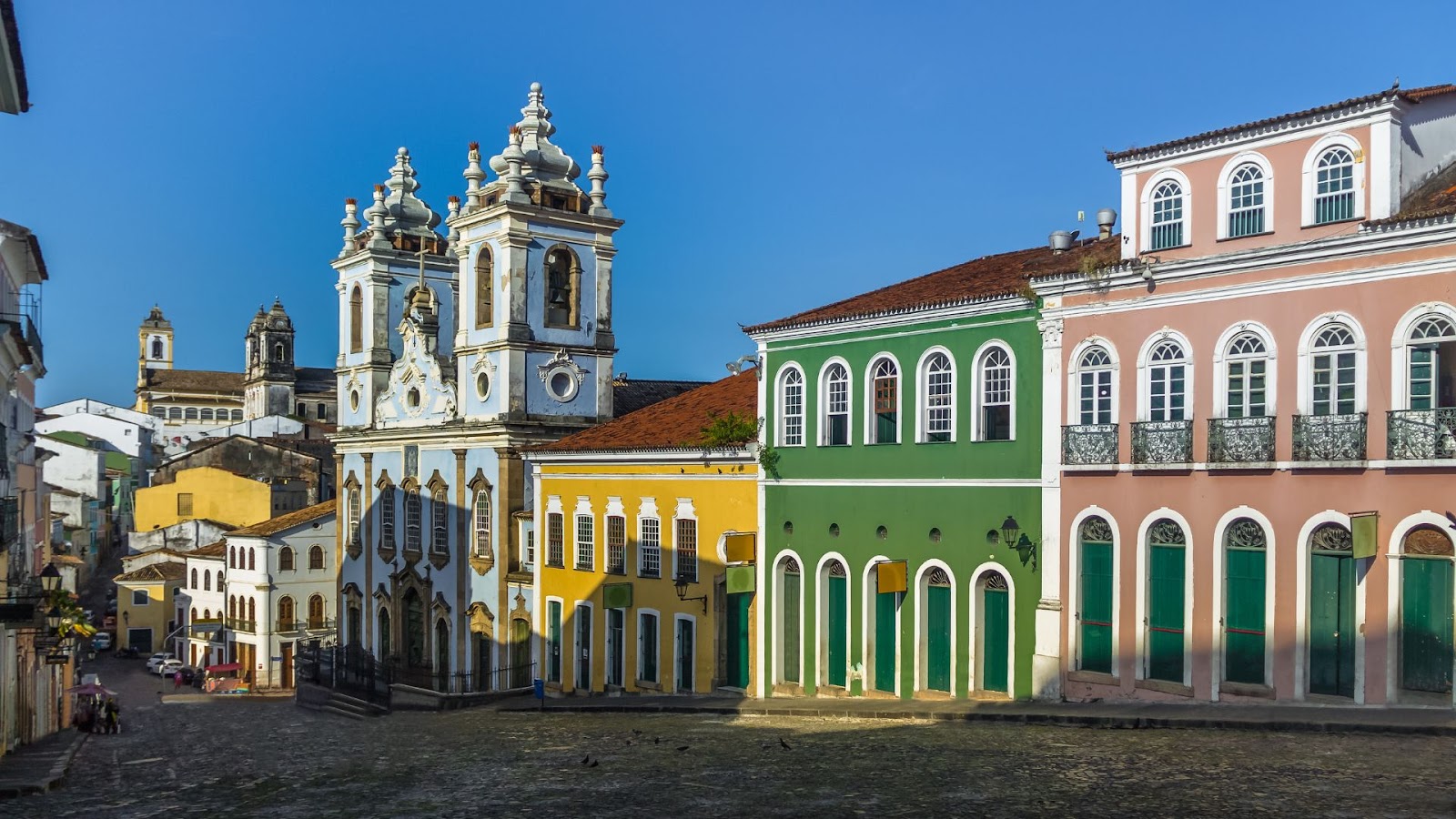 Arquitetura em Salvador Uma paisagem urbana vibrante do Centro Histórico de Salvador, Pelourinho, sob um céu azul claro. A imagem mostra uma rua de paralelepípedos ladeada por edifícios coloniais coloridos. Em destaque, à esquerda, está uma igreja barroca com fachadas ornamentadas em tons de azul claro e branco e duas torres. Ao lado da igreja, há uma série de casas com fachadas em amarelo, verde vibrante e rosa antigo, todas com janelas brancas e detalhes arquitetônicos clássicos. Ao fundo, outras construções históricas podem ser avistadas, completando a atmosfera característica do local.