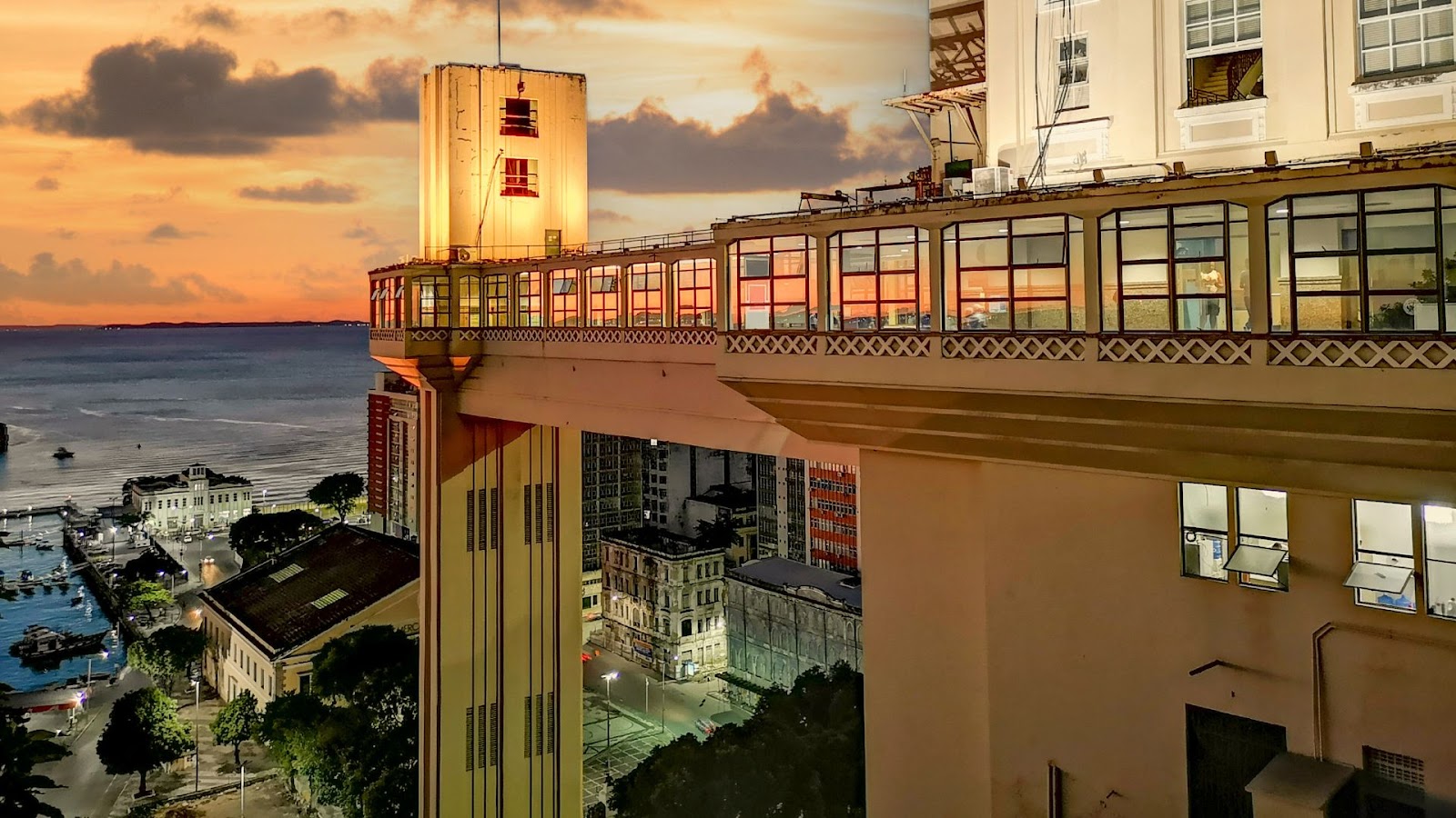 Arquitetura em Salvador Uma vista impressionante do Elevador Lacerda em Salvador, Bahia, ao pôr do sol. A estrutura imponente do elevador, com suas torres iluminadas em tons quentes, domina o centro da imagem. As passarelas e a parte superior da estrutura, com janelas envidraçadas, refletem as cores vibrantes do céu alaranjado e roxo do entardecer. Ao fundo, a Baía de Todos-os-Santos se estende, com alguns barcos e a orla da cidade visíveis. Abaixo do elevador, parte da cidade baixa com seus edifícios e iluminação noturna começa a aparecer.